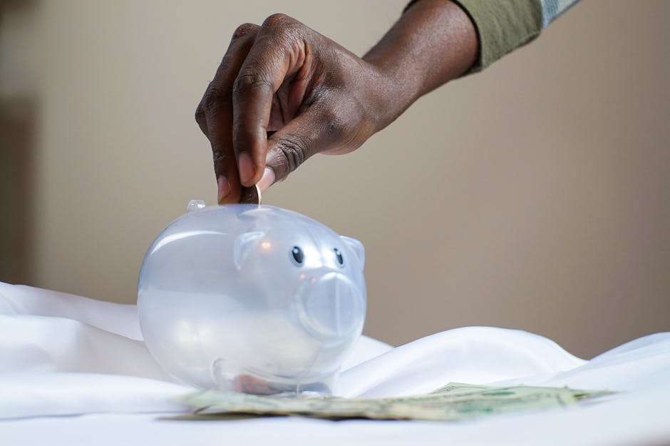 Close-up of a person's hand placing coins into a transparent piggy bank to save money